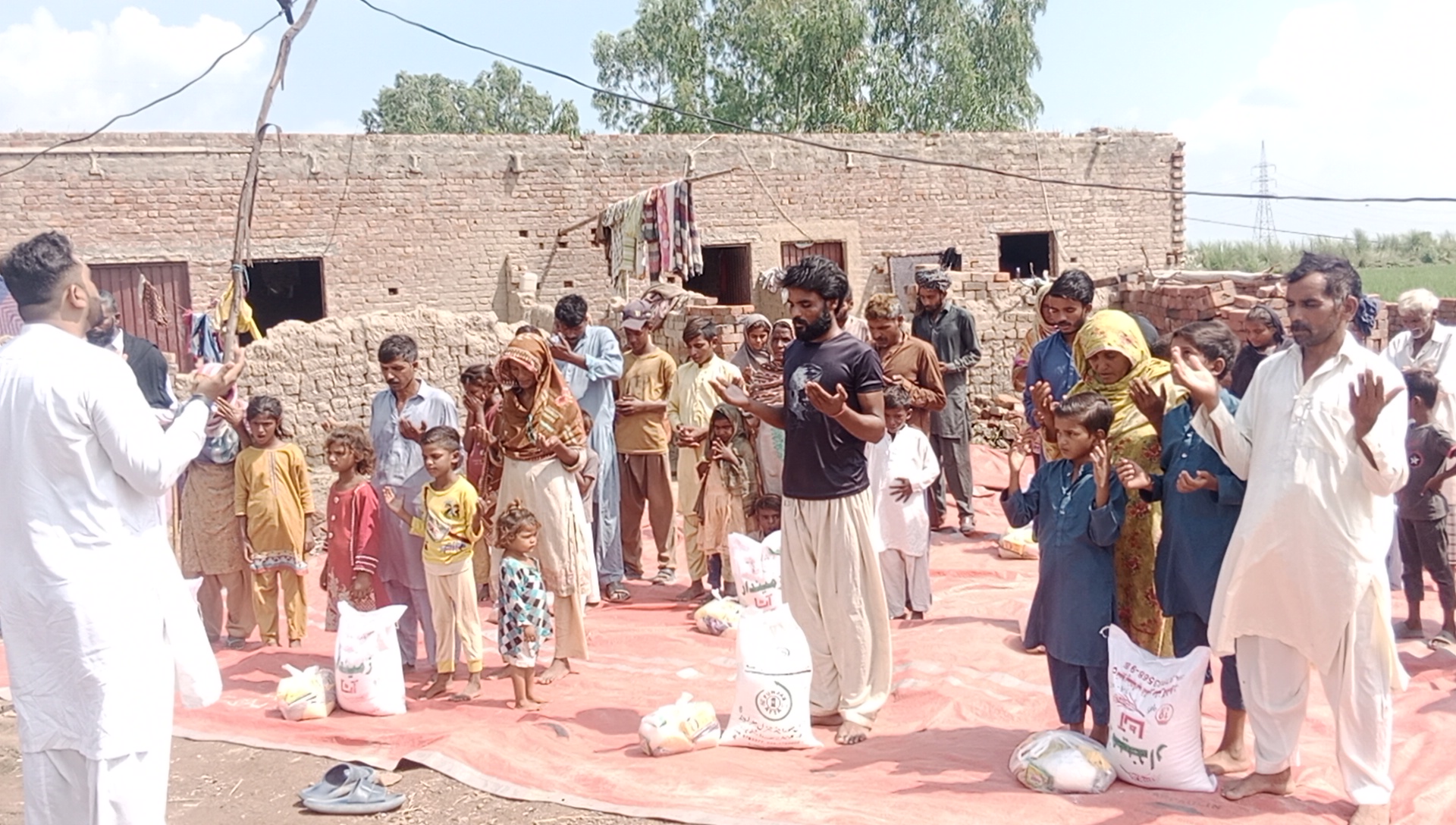 Flood-affected brick kiln families standing in prayer around bags of flour and food supplies